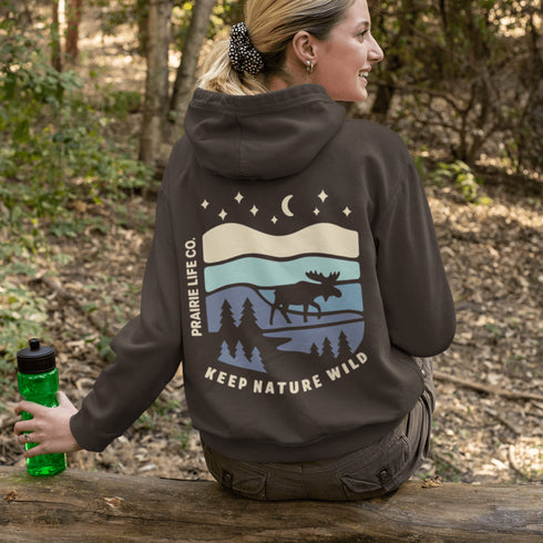 This hoodie features a moose silhouette roaming across prairie hills, pine trees with stars scattered above and a crescent moon glowing softly.