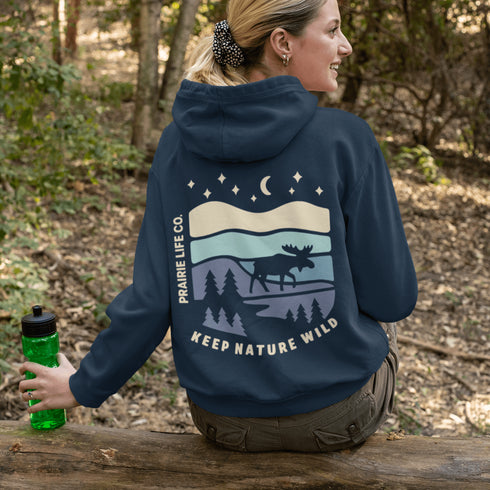 This hoodie features a moose silhouette roaming across prairie hills, pine trees with stars scattered above and a crescent moon glowing softly.