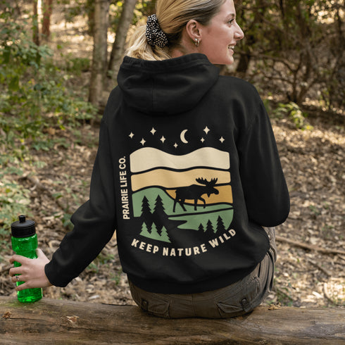 This premium hoodie features a moose silhouette wandering across prairie hills, Pine trees and a clear night sky filled with stars and a crescent moon.