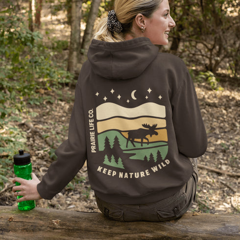 This premium hoodie features a moose silhouette wandering across prairie hills, Pine trees and a clear night sky filled with stars and a crescent moon.
