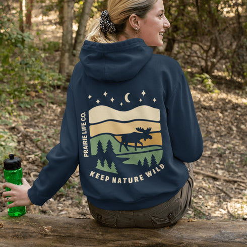This premium hoodie features a moose silhouette wandering across prairie hills, Pine trees and a clear night sky filled with stars and a crescent moon.