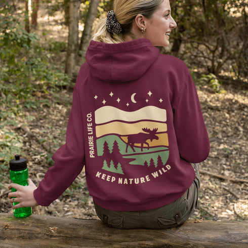 This premium hoodie features a moose silhouette wandering across prairie hills, Pine trees and a clear night sky filled with stars and a crescent moon.