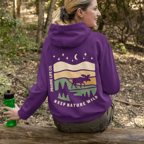 This premium hoodie features a moose silhouette wandering across prairie hills, Pine trees and a clear night sky filled with stars and a crescent moon.
