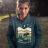 Featuring a bold moose silhouette layered over rolling hills, pine trees, and a starry night sky, this hoodie is a tribute to Canada’s wild landscapes.