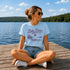 Woman wearing a light blue Lake Days graphic t-shirt while sitting on a dock overlooking a calm lake on a sunny summer day.