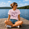 Woman wearing a pink Lake Days graphic t-shirt while sitting on a dock overlooking a calm lake on a sunny summer day.