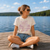 Woman sitting on a dock by a lake wearing a t-shirt with text and sunglasses.