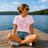 Woman sitting on a dock by a lake wearing a pink t-shirt with purple text and graphics.