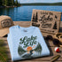 Stack of 'Lake Life' t-shirts with a cap, sunglasses, and wooden sign by a lake.