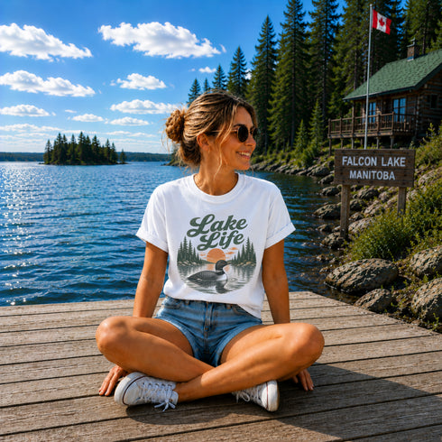 Woman sitting on a dock by a lake wearing a 'Lake Life' t-shirt with a cabin and flag in the background.