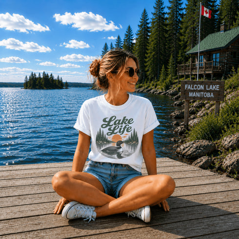 Woman sitting on a dock by a lake wearing a 'Lake Life' t-shirt with a cabin and flag in the background.