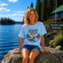 Woman wearing a 'Lake Life' t-shirt sitting on a rock by a lake with trees and a cabin in the background.