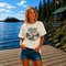 Woman wearing a vintage Lake Life loon graphic t-shirt while standing on a dock overlooking Falcon Lake, Manitoba with pine trees, a lakeside cabin, and a peaceful Canadian lake in the background.