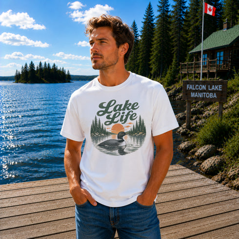 Man wearing a vintage Lake Life loon graphic t-shirt while standing on a dock overlooking Falcon Lake, Manitoba with pine trees, a lakeside cabin, and a peaceful Canadian lake in the background.