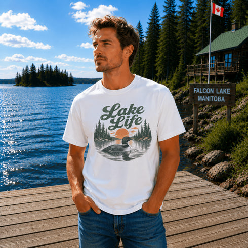 Man wearing a vintage Lake Life loon graphic t-shirt while standing on a dock overlooking Falcon Lake, Manitoba with pine trees, a lakeside cabin, and a peaceful Canadian lake in the background.