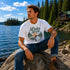 Man sitting on a rock by a lake wearing a 'Lake Life' t-shirt with a scenic design.