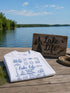 White t-shirt with 'Lake Life' design on a wooden dock by a lake.