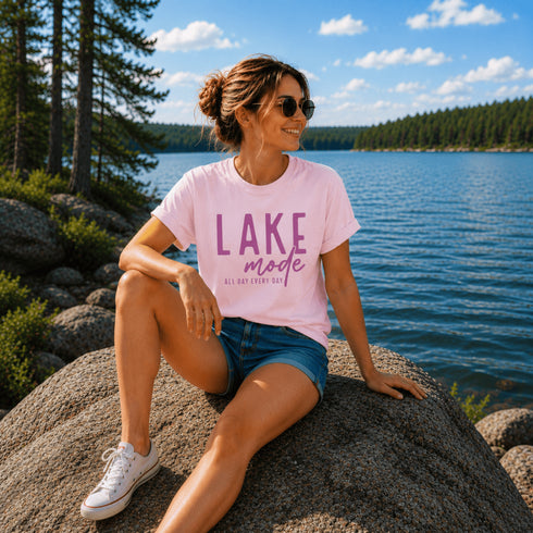 Woman sitting by a lake wearing a pink 'Lake Mode' t-shirt.