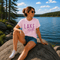 Woman sitting by a lake wearing a pink 'Lake Mode' t-shirt.