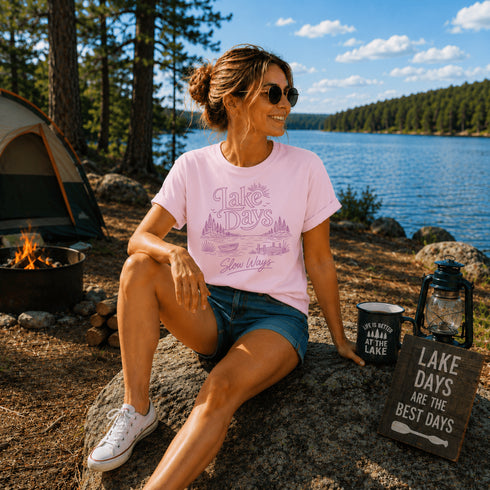 Woman sitting by a lake with a pink t-shirt and 'Lake Days' mug.