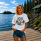 Woman wearing a vintage Lake Life loon graphic t-shirt while standing on a dock overlooking Falcon Lake, Manitoba with pine trees, a lakeside cabin, and a peaceful Canadian lake in the background.