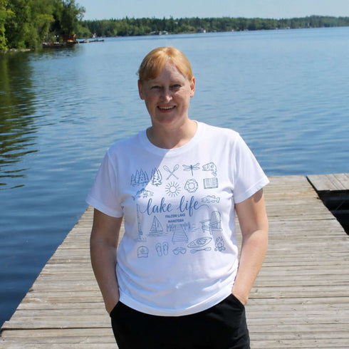 Person wearing a white t-shirt with graphics and text, standing on a wooden dock by a lake.