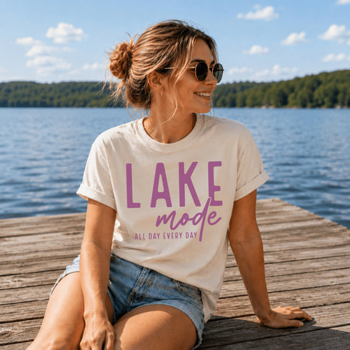 Woman sitting on a dock by a lake wearing a 'Lake Mode' t-shirt.