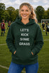 Woman wearing a green hoodie with 'LET'S KICK SOME GRASS' text on a soccer field.