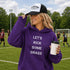 Woman on a soccer field wearing a purple hoodie with text and a black and white cap.