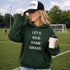 Woman on a soccer field wearing a green hoodie with text and a cap, holding a coffee cup.