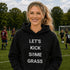 Woman wearing a black hoodie with 'LET'S KICK SOME GRASS' text on a soccer field.