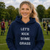 Woman wearing a navy hoodie with 'LET'S KICK SOME GRASS' text on a soccer field.