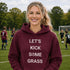 Woman wearing a maroon hoodie with 'Let's Kick Some Grass' text on a soccer field.
