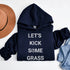 Navy blue hoodie with 'Let's Kick Some Grass' text, placed on a white surface with a hat and denim jacket.