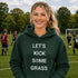 Woman wearing “Let’s Kick Some Grass” hoodie standing on a soccer field during a youth soccer game