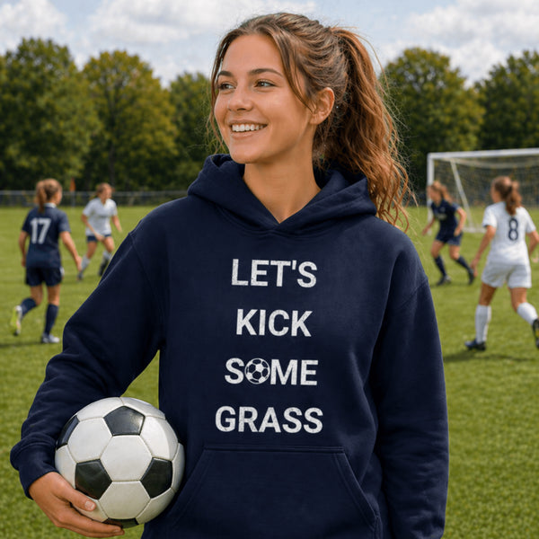Person wearing a navy hoodie with 'LET'S KICK SOME GRASS' text, holding a soccer ball on a soccer field.
