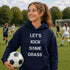 Person wearing a navy hoodie with 'LET'S KICK SOME GRASS' text, holding a soccer ball on a soccer field.