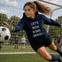 Soccer goalkeeper in a navy hoodie with text, diving for a ball on a soccer field.