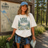 Woman wearing the Lost Among the Pines at Golden Hour hiking t-shirt featuring a vintage mountain and forest landscape design while standing beside a Banff Canada trail map in a forest setting.