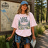 Woman wearing the Lost Among the Pines at Golden Hour hiking t-shirt featuring a vintage mountain and forest landscape design while standing beside a Banff Canada trail map in a forest setting.
