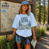 Woman wearing the Lost Among the Pines at Golden Hour hiking t-shirt featuring a vintage mountain and forest landscape design while standing beside a Banff Canada trail map in a forest setting.