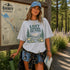Woman wearing the Lost Among the Pines at Golden Hour hiking t-shirt featuring a vintage mountain and forest landscape design while standing beside a Banff Canada trail map in a forest setting.
