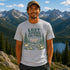 Man wearing a t-shirt with a nature graphic and text, standing in front of mountains and a lake.