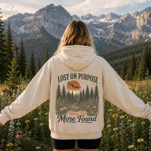 A woman stands in a mountain meadow with her back to the camera, wearing a cream-colored hoodie with a large forest graphic and the text 'LOST ON PURPOSE Never Felt More Found' printed on the back. Snow-capped mountains and pine trees are visible in the background.