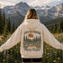 A woman stands in a mountain meadow with her back to the camera, wearing a cream-colored hoodie with a large forest graphic and the text 'LOST ON PURPOSE Never Felt More Found' printed on the back. Snow-capped mountains and pine trees are visible in the background.