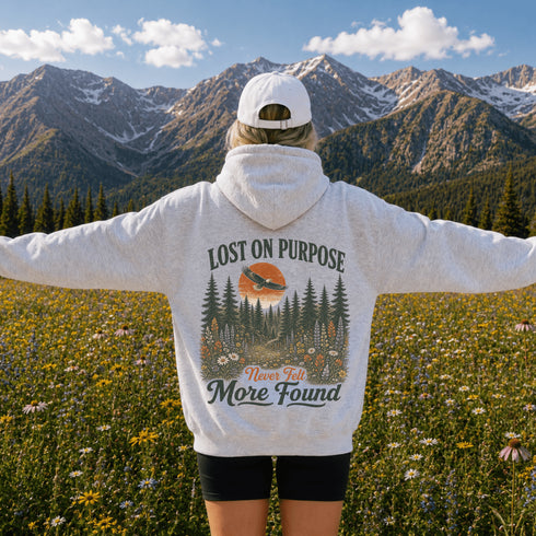 A person standing in a mountain meadow with arms outstretched, wearing a light heather gray hoodie with a large back graphic that reads 'LOST ON PURPOSE Never Felt More Found' above a forest and sunset scene, plus a white cap and black shorts.