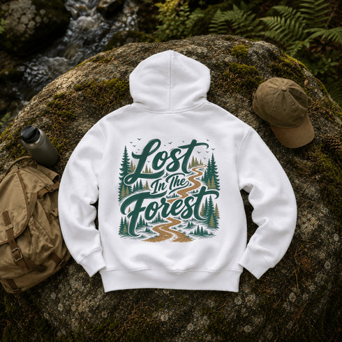 A white pullover hoodie laid flat on a mossy rock outdoors, showing a large back graphic that reads “Lost in the Forest” with trees and a winding path. A tan cap, backpack, and water bottle are placed around it, with a forest stream and ferns in the background.