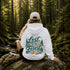 A person sitting on a mossy log in a forest, viewed from behind, wearing a white hoodie with a large graphic on the back that reads 'Lost in the Forest' in green and brown lettering with pine trees. A beige cap, hiking boots, and a backpack are beside them.