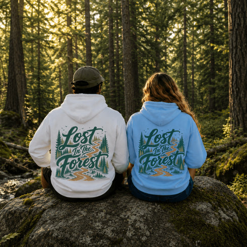 Two people sitting on a mossy rock in a sunlit forest, seen from behind, wearing matching hoodies with a large scenic graphic that reads 'Lost in the Forest' in teal and orange lettering with pine trees and a winding path.