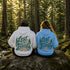 Two people sitting on a mossy rock in a sunlit forest, seen from behind, wearing matching hoodies with a large scenic graphic that reads 'Lost in the Forest' in teal and orange lettering with pine trees and a winding path.